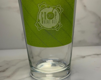 May include: A clear pint glass with a green label featuring the text "Crop Wheat Ale" in a stylized design. The glass is empty and reflects light, suggesting it's ready for a beverage.