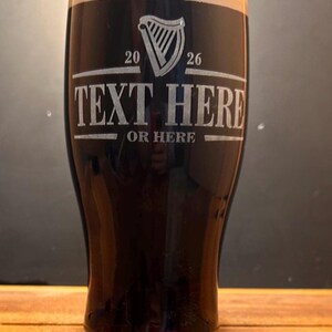 May include: A tall, dark beer glass filled with a stout, topped with a creamy head. The glass features etched text: "TEXT HERE OR HERE" and a harp symbol with the numbers "20" and "26". The glass sits on a wooden surface.