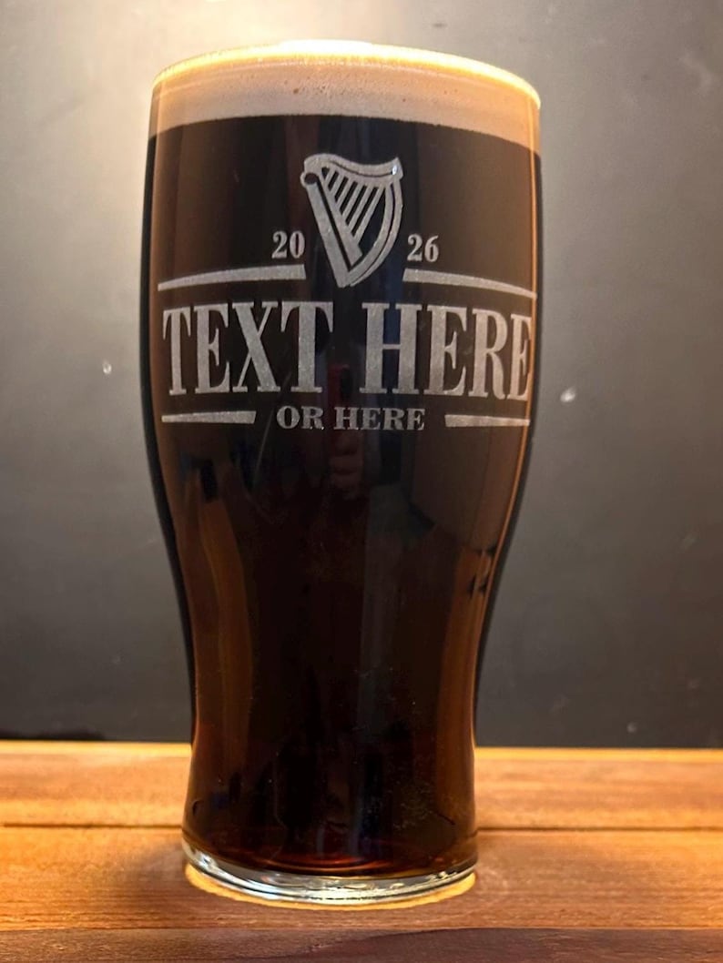 May include: A tall, dark beer glass filled with a stout, topped with a creamy head. The glass features etched text: "TEXT HERE OR HERE" and a harp symbol with the numbers "20" and "26". The glass sits on a wooden surface.