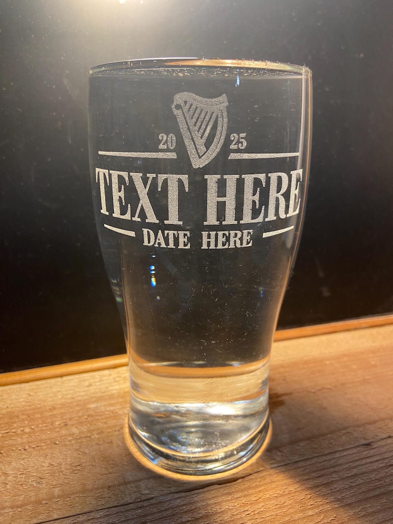 May include: A clear glass pint glass with an etched design of a harp, the numbers 20 and 25, the text "TEXT HERE" and "DATE HERE".
