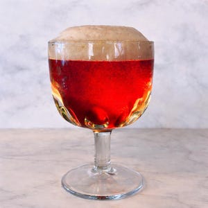 May include: A clear, stemmed glass filled with a reddish-brown beverage, topped with a thick, tan foam. The glass has a decorative, textured design and sits on a marble surface. The drink appears to be beer.