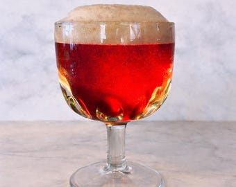 May include: A clear, stemmed glass filled with a reddish-brown beverage, topped with a thick, tan foam. The glass has a decorative, textured design and sits on a marble surface. The drink appears to be beer.