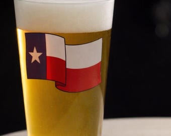 May include: A pint glass filled with beer and topped with a foamy head. The glass features the Texas flag on the front. The beer is a light amber color, and the background is blurred.