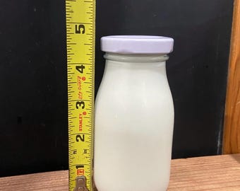 May include: A clear glass milk bottle filled with white liquid, topped with a light purple lid. The bottle is approximately 5 inches tall, as measured by a yellow tape measure. The background is a dark chalkboard and wooden surface.