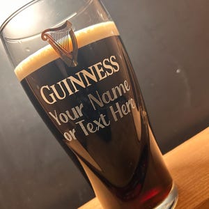 May include: A pint glass filled with dark stout beer and a creamy head. The glass features the Guinness logo and the text "Your Name or Text Here". The glass is angled, showcasing the beer's colour and the custom text.