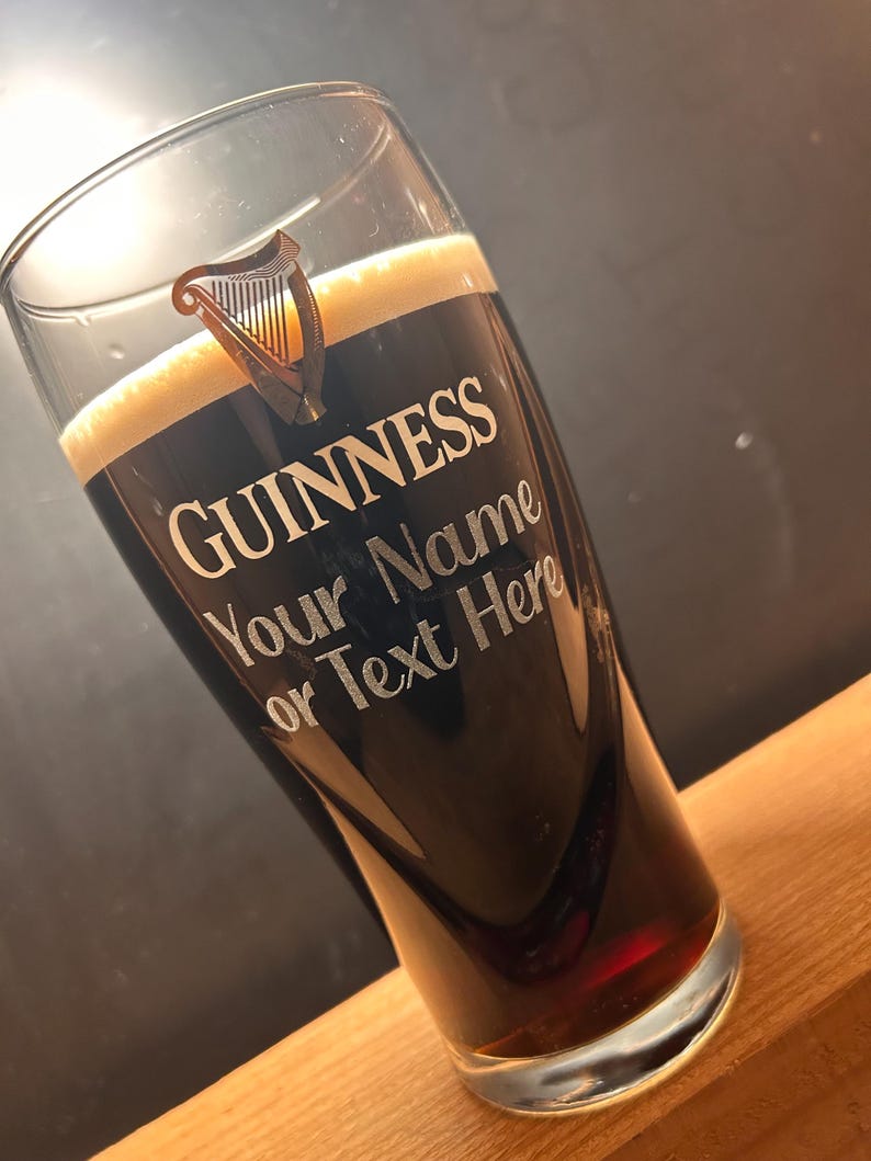 May include: A pint glass filled with dark stout beer and a creamy head. The glass features the Guinness logo and the text "Your Name or Text Here". The glass is angled, showcasing the beer's colour and the custom text.