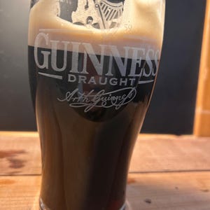 May include: A pint glass of Guinness Draught, a dark stout with a creamy head. The Guinness logo and Arthur Guinness' signature are etched on the glass. The glass is set on a wooden surface. The beer is a deep, rich colour.