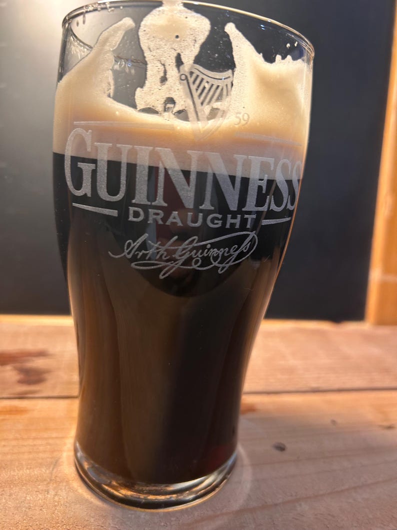 May include: A pint glass of Guinness Draught, a dark stout with a creamy head. The Guinness logo and Arthur Guinness' signature are etched on the glass. The glass is set on a wooden surface. The beer is a deep, rich colour.