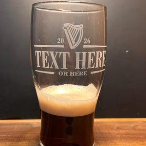May include: A clear pint glass filled with dark beer and a foamy head. The glass has etched text that reads "TEXT HERE OR HERE" with a harp symbol and the numbers "20 26". The glass is on a wooden surface.