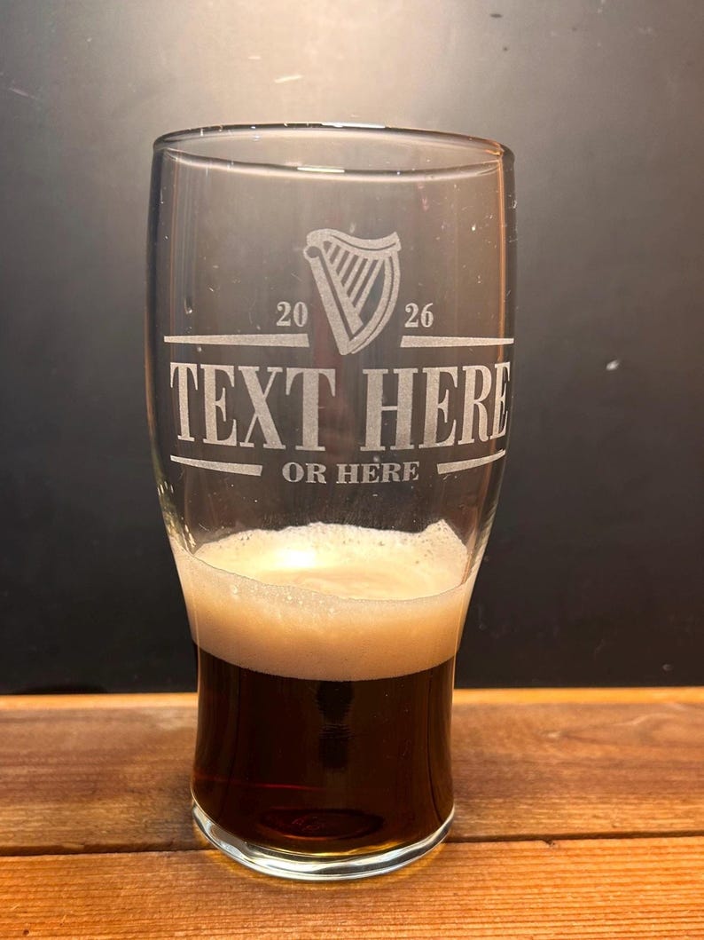 May include: A clear pint glass filled with dark beer and a foamy head. The glass has etched text that reads "TEXT HERE OR HERE" with a harp symbol and the numbers "20 26". The glass is on a wooden surface.