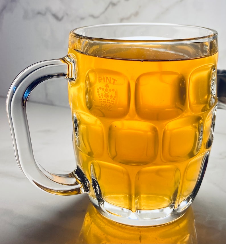 K&ouml;nnte beinhalten: Ein klares Glas-Bierglas mit einer strukturierten Oberfl&auml;che und dem Wort "PINT" auf der Seite. Das Glas ist mit einer hellgelben Fl&uuml;ssigkeit gef&uuml;llt.