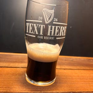May include: A clear pint glass with a dark stout beer and a foamy head. The glass has etched text that reads "TEXT HERE" and "OR HERE" with a harp symbol and the numbers "20" and "26". The glass sits on a wooden surface.