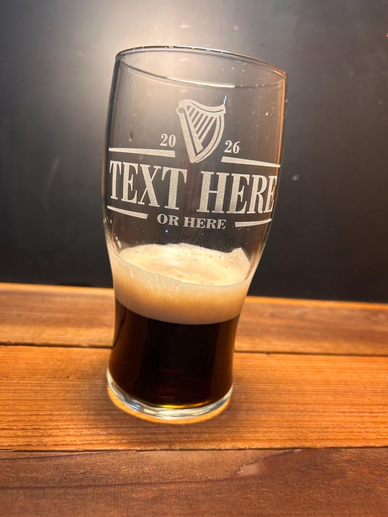 May include: A clear pint glass with a dark stout beer and a foamy head. The glass has etched text that reads "TEXT HERE" and "OR HERE" with a harp symbol and the numbers "20" and "26". The glass sits on a wooden surface.
