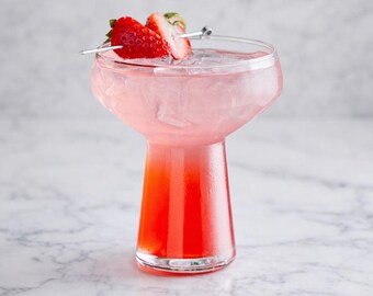 May include: A refreshing pink cocktail in a unique glass, garnished with fresh strawberries on a skewer. The drink has a gradient of red at the bottom, fading to pink. The glass sits on a marble surface.