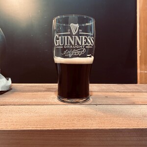 May include: A pint glass of Guinness Draught, filled with dark stout and a creamy white head. The glass features the Guinness logo and the words "Guinness Draught" in white lettering. The glass is set on a wooden surface.