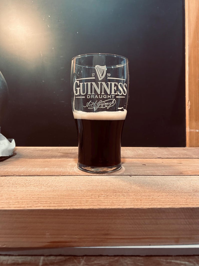 May include: A pint glass of Guinness Draught, filled with dark stout and a creamy white head. The glass features the Guinness logo and the words "Guinness Draught" in white lettering. The glass is set on a wooden surface.