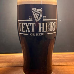 May include: A tall pint glass filled with dark stout and a creamy head. The glass features an etched design with a harp symbol, the numbers 20 and 26, and the words "TEXT HERE" and "OR HERE". The glass is set on a wooden surface.