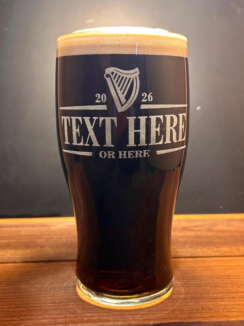 May include: A tall pint glass filled with dark stout and a creamy head. The glass features an etched design with a harp symbol, the numbers 20 and 26, and the words "TEXT HERE" and "OR HERE". The glass is set on a wooden surface.