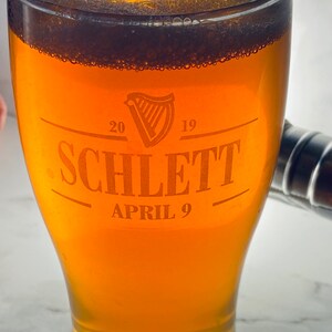 May include: A glass of beer with a white head in a clear glass pint glass. The glass has an etched design that says "Schlett" with the years "20" and "19" and the date "April 9".