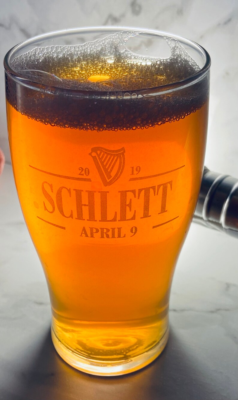 May include: A glass of beer with a white head in a clear glass pint glass. The glass has an etched design that says "Schlett" with the years "20" and "19" and the date "April 9".