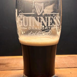 May include: A pint glass of Guinness Draught stout. The glass is etched with the Guinness logo, the words "Guinness Draught" and "Arth Guinness". The stout is a dark brown colour with a creamy, tan head.