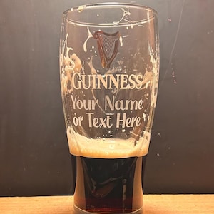 May include: A clear pint glass filled with dark stout and a creamy head. The glass is etched with the Guinness logo and the text "Your Name or Text Here". The glass sits on a wooden surface.