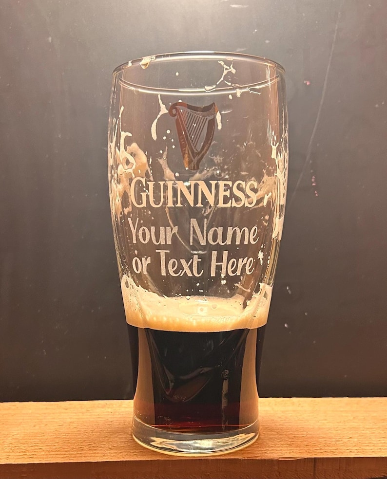 May include: A clear pint glass filled with dark stout and a creamy head. The glass is etched with the Guinness logo and the text "Your Name or Text Here". The glass sits on a wooden surface.