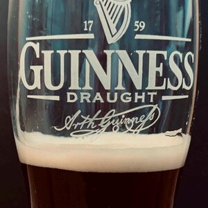 May include: A pint glass filled with dark stout, topped with a creamy white head. The glass features the Guinness logo, including a harp symbol and the words "Guinness Draught". The text "1759" is also visible.