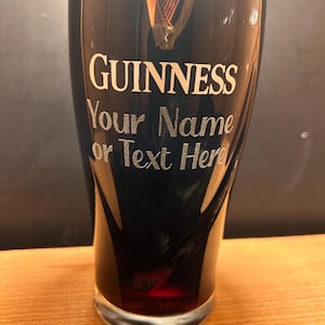 May include: A pint glass filled with a dark beverage, likely Guinness. The glass has the Guinness logo and the text "GUINNESS Your Name or Text Here" in white. The glass sits on a wooden surface.
