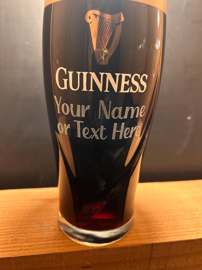 May include: A pint glass filled with a dark beverage, likely Guinness. The glass has the Guinness logo and the text "GUINNESS Your Name or Text Here" in white. The glass sits on a wooden surface.