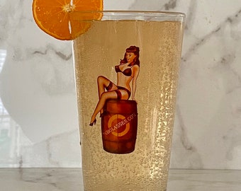 May include: A tall glass filled with a light-colored beverage, garnished with a slice of orange. The glass has a graphic of a woman sitting on a barrel with the text "OBGLASSES.CO."