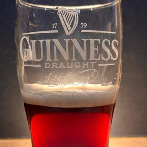 May include: A pint glass of Guinness draught beer. The glass is etched with the Guinness logo, the year 1759, and Arthur Guinness' signature. The beer is a dark red colour with a thick, creamy white head.