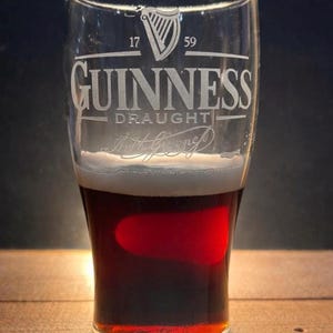 May include: A pint glass of Guinness stout, with a creamy head and a dark, reddish-brown colour. The glass is etched with the Guinness logo, a harp, and the text "GUINNESS DRAUGHT". The background is dark.