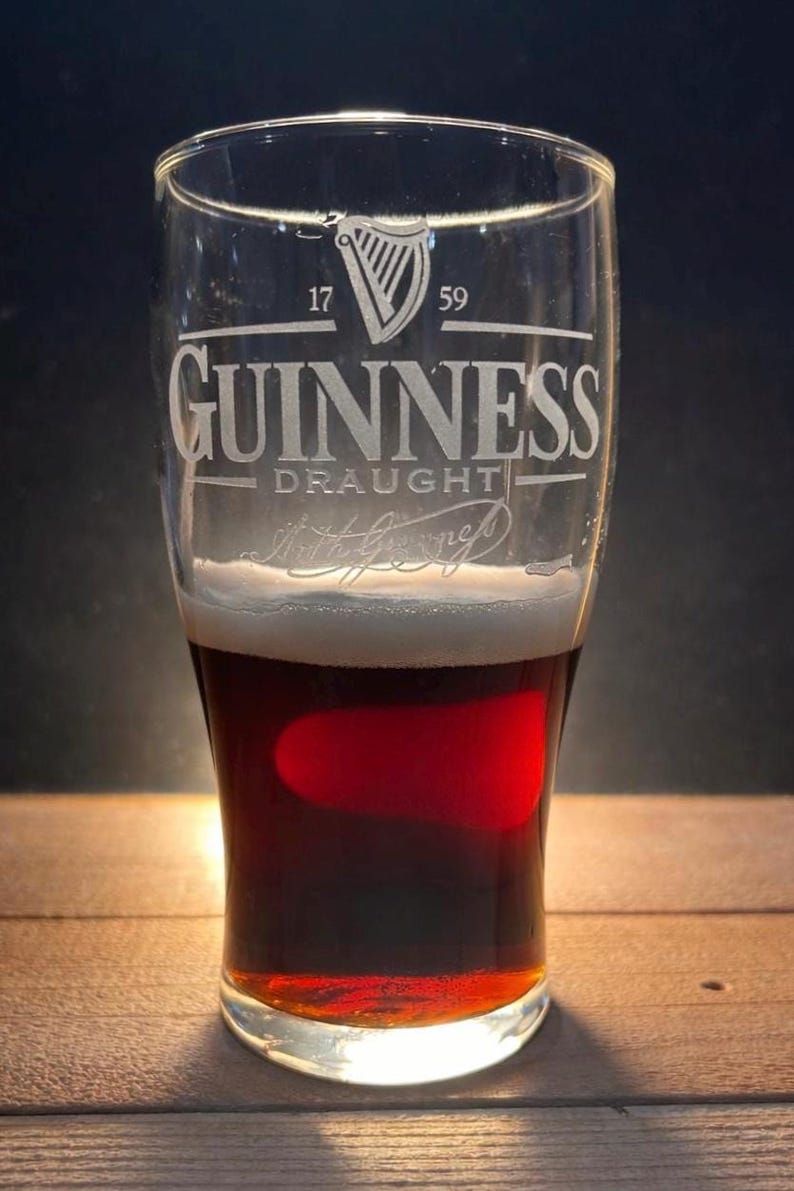 May include: A pint glass of Guinness stout, with a creamy head and a dark, reddish-brown colour. The glass is etched with the Guinness logo, a harp, and the text "GUINNESS DRAUGHT". The background is dark.