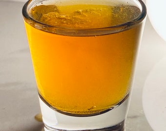 May include: A clear glass shot glass filled with a golden yellow liquid. The liquid has a slight head of foam on top.