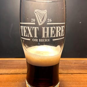 May include: A clear pint glass with a dark stout and a foamy head. The glass has etched text that reads "TEXT HERE OR HERE" with a harp symbol and the numbers "20 26". The glass is on a wooden surface.