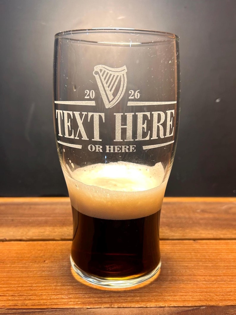 May include: A clear pint glass with a dark stout and a foamy head. The glass has etched text that reads "TEXT HERE OR HERE" with a harp symbol and the numbers "20 26". The glass is on a wooden surface.