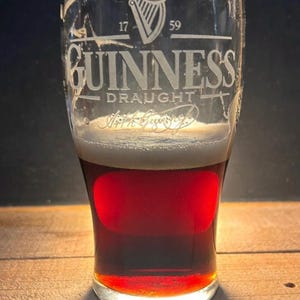 May include: A pint glass of Guinness stout, displaying the brand's logo and the year 1759. The beer has a creamy head and a dark, reddish-brown body. The glass sits on a wooden surface.
