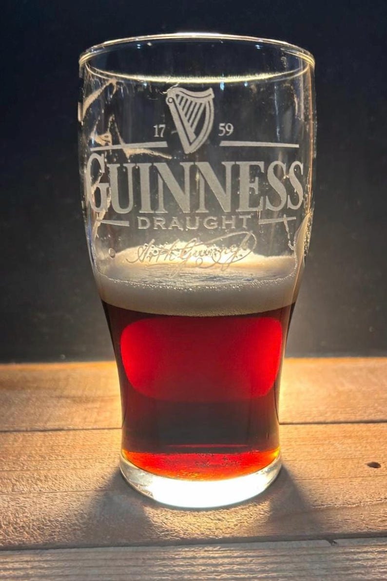 May include: A pint glass of Guinness stout, displaying the brand's logo and the year 1759. The beer has a creamy head and a dark, reddish-brown body. The glass sits on a wooden surface.