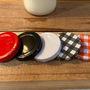 May include: A collection of jar lids in various colors and patterns. The lids include solid red, black, white, black and white checkered, and orange and white checkered designs. The lids are arranged on a wooden surface.