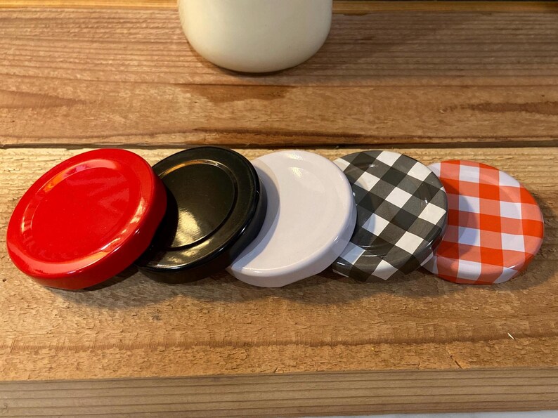 May include: A collection of jar lids in various colors and patterns. The lids include solid red, black, white, black and white checkered, and orange and white checkered designs. The lids are arranged on a wooden surface.