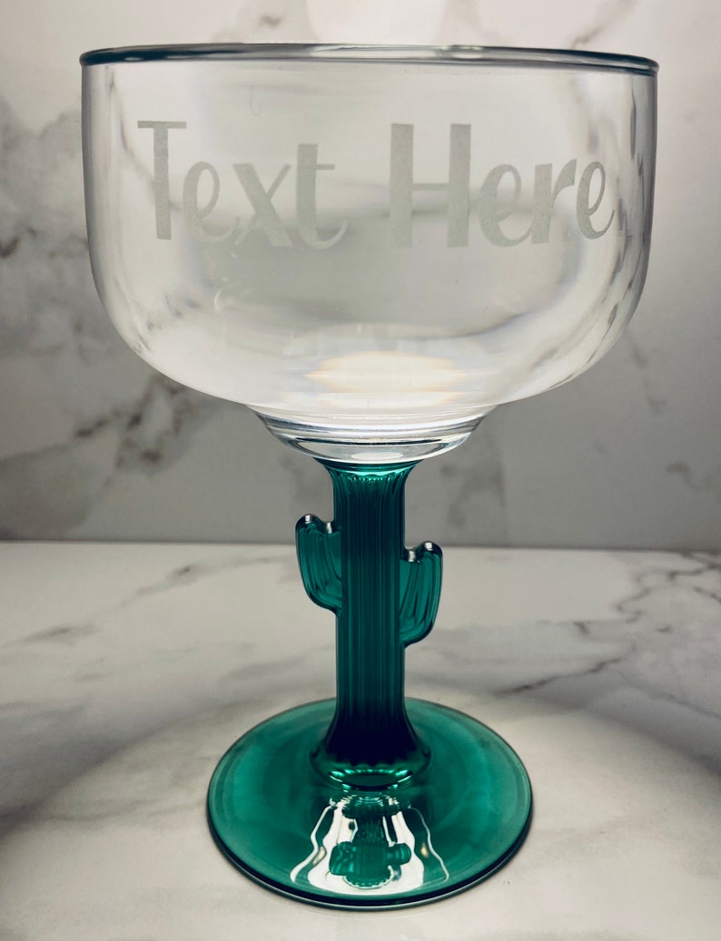 May include: Clear margarita glass with a green cactus stem and base. The text "Text Here" is etched on the bowl of the glass. The glass is designed for cocktails and beverages.