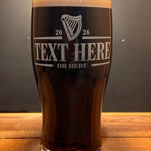 May include: A tall glass filled with a dark stout, topped with a creamy foam. The glass features an etched design with a harp symbol, the numbers "20" and "26", and the text "TEXT HERE" and "OR HERE". The glass sits on a wooden surface.