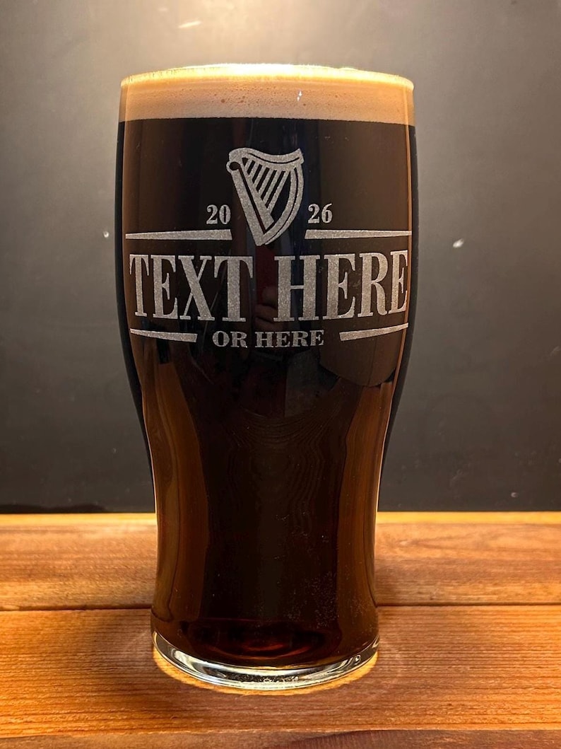 May include: A tall glass filled with a dark stout, topped with a creamy foam. The glass features an etched design with a harp symbol, the numbers "20" and "26", and the text "TEXT HERE" and "OR HERE". The glass sits on a wooden surface.