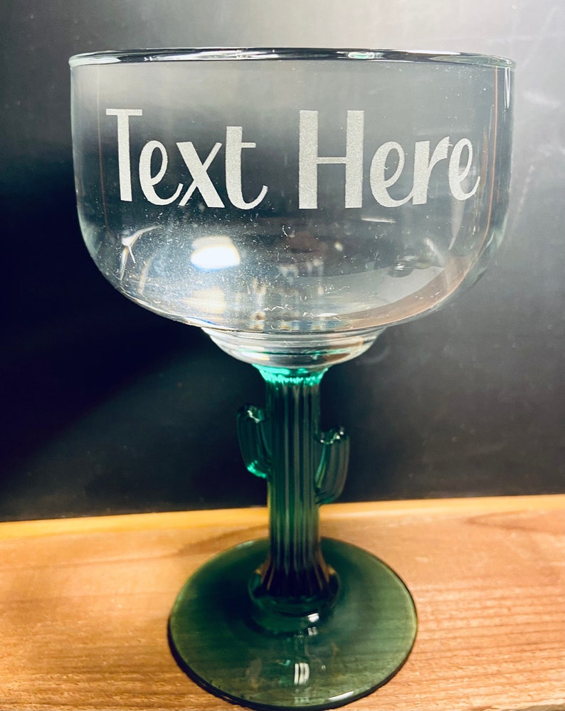 May include: A clear margarita glass with a green cactus-shaped stem and base. The glass has the words "Text Here" etched on the bowl. The glass is designed for cocktails and beverages.