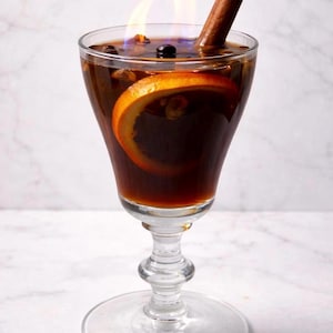 May include: A stemmed glass filled with a dark brown liquid, an orange slice, and a cinnamon stick. Blue flames rise from the drink, suggesting a cocktail or beverage with a fiery presentation. The glass sits on a white surface.