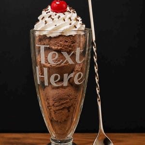 May include: A tall glass sundae dish filled with scoops of brown chocolate ice cream, topped with white whipped cream, chocolate sprinkles, and a bright red cherry. The glass features the etched text "Text Here" and sits beside a long metal spoon.