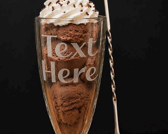 May include: A tall glass sundae dish filled with scoops of brown chocolate ice cream, topped with white whipped cream, chocolate sprinkles, and a bright red cherry. The glass features the etched text "Text Here" and sits beside a long metal spoon.