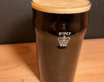 May include: A clear glass pint beer glass filled with a dark brown stout topped with a thick tan foam head. The glass features a white etched crown logo above the text PINT and the number 562 printed on the front surface of the glass.