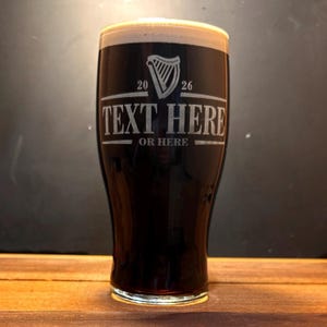 May include: A pint glass filled with dark beer and a creamy head. The glass has etched text that reads "TEXT HERE" and "OR HERE" below a harp symbol and the numbers "20" and "26". The glass is on a wooden surface.